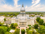 Illinois budget bill targets sports prediction markets with unique transaction tax Aerial view of the Illinois State Capitol building in Springfield, Illinois, showcasing the historic government landmark, iconic dome, landscaped grounds, and downtown skyline under a bright blue sky. Illinois budget bill targets sports prediction markets with unique transaction tax